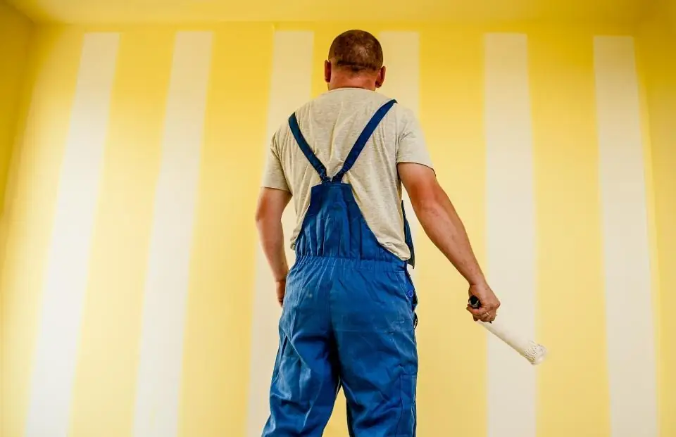 Peintre en salopette, rouleau à la main, devant un mur à rayures jaunes et blanches
