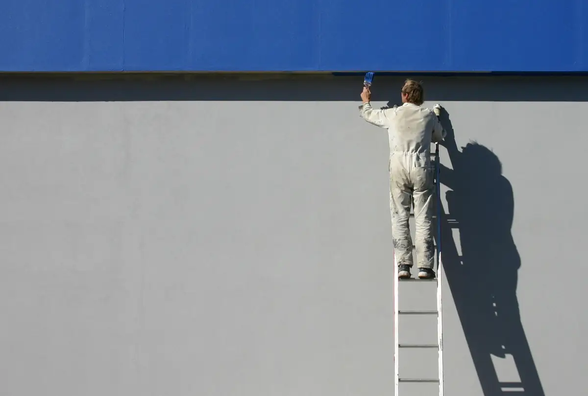 Peintre en bâtiment sur une échelle, qui reprend le haut d’un grand mur gris