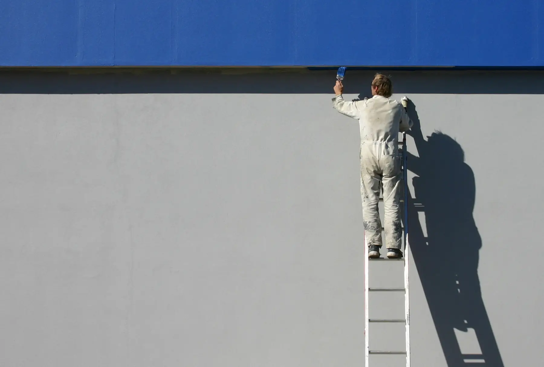 Peintre en bâtiment sur une échelle, qui reprend le haut d’un grand mur gris