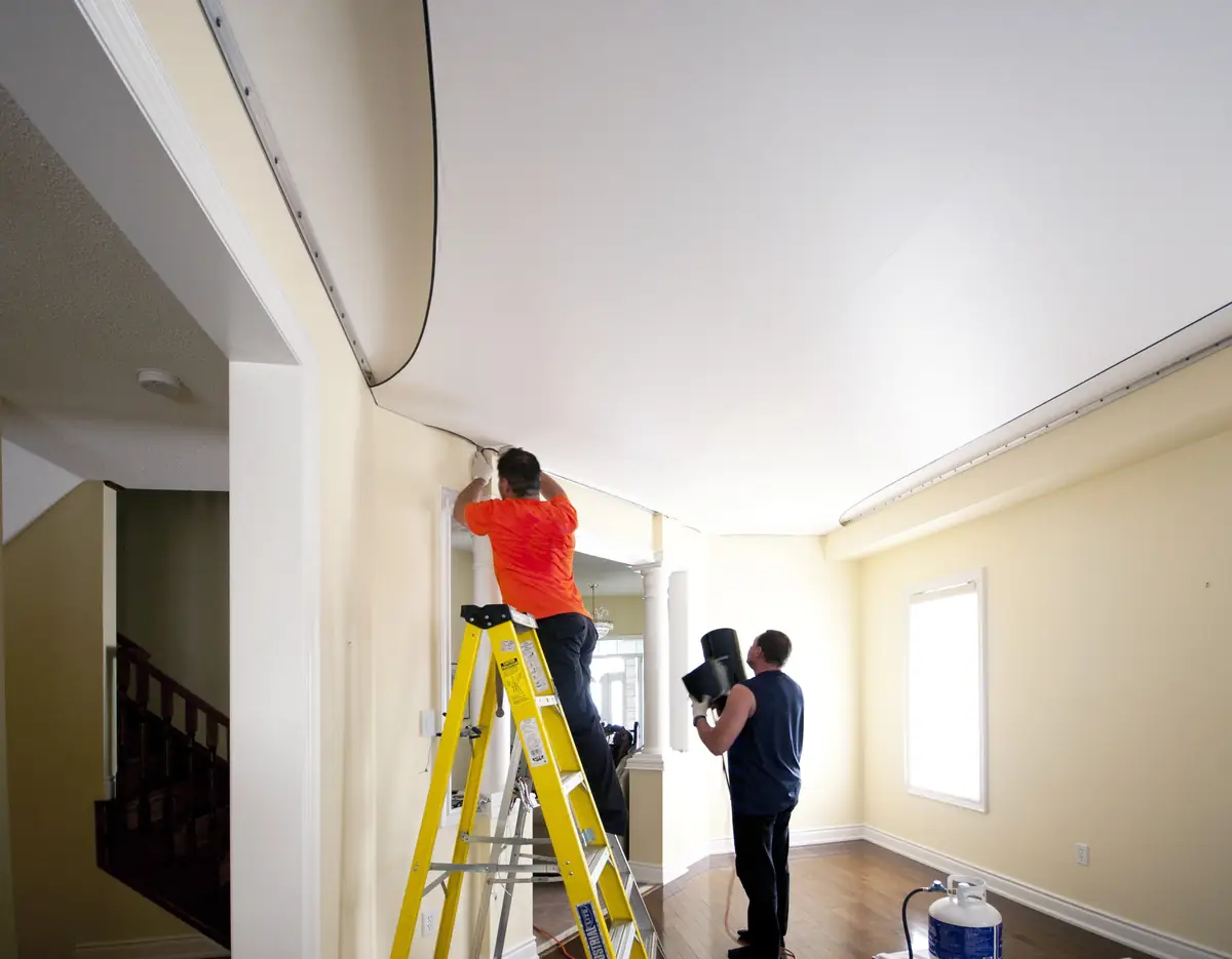 Deux poseurs installent une toile de plafond tendu dans une pièce vide, l’un sur un escabeau