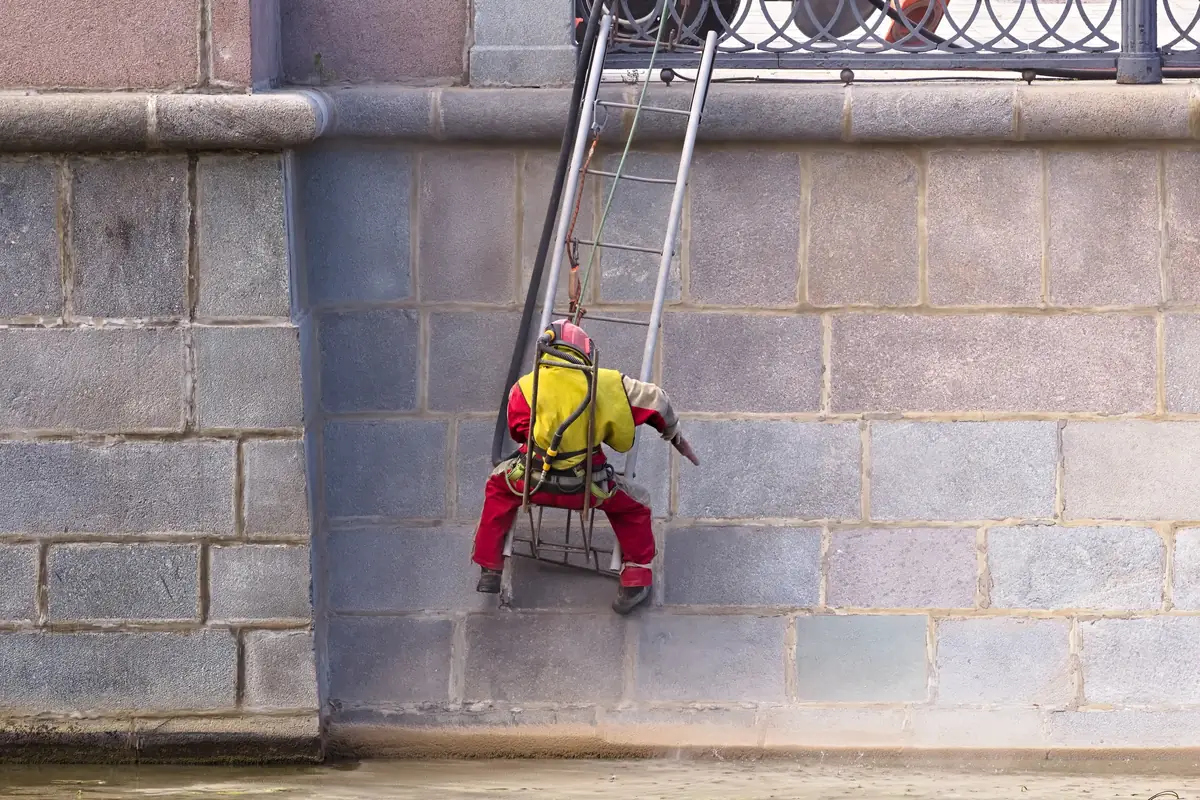 Ouvrier suspendu qui sable un parement de granit, équipé d’une combinaison et d’un casque ventilé