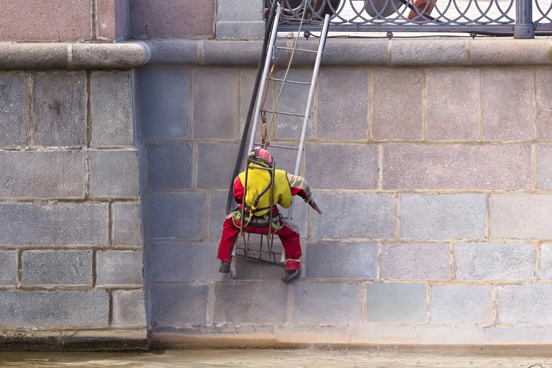 Ouvrier suspendu qui sable un parement de granit, équipé d’une combinaison et d’un casque ventilé